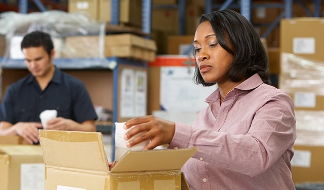 A woman in the foreground packing items into a box with a male colleague in the background organizing inventory for retail brand fulfillment