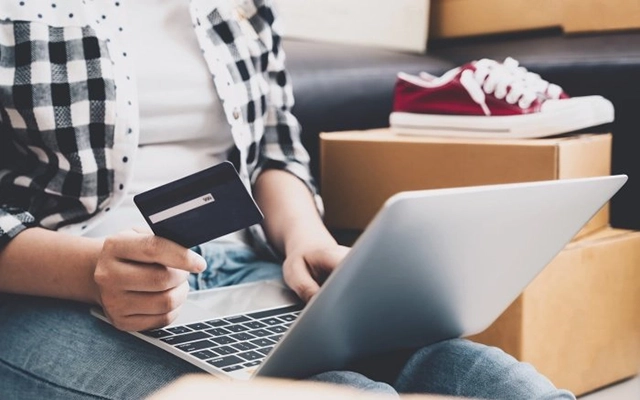Woman sitting on couch wearing a plaid shirt and jeans places an order on a laptop with a credit card while boxes and shoes sit beside her